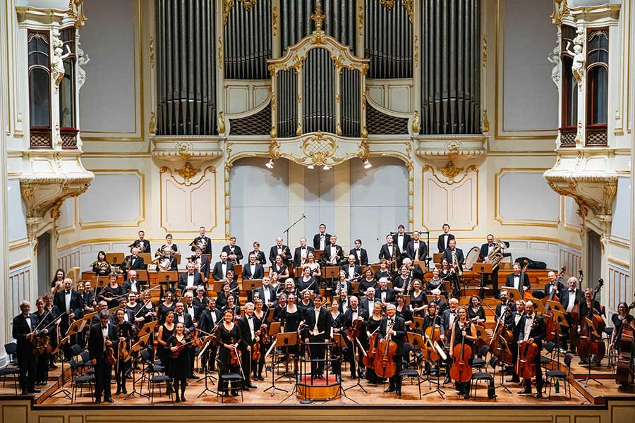 Großes Sinfonieorchester in Konzertkleidung auf einer Bühne vor einer prunkvollen weißen Orgel mit goldenen Verzierungen.