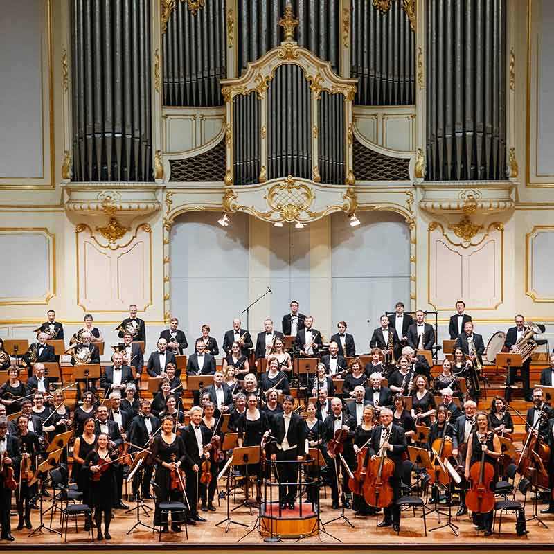 Großes Sinfonieorchester in Konzertkleidung auf einer Bühne vor einer prunkvollen weißen Orgel mit goldenen Verzierungen.